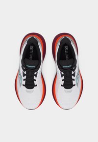 Ζευγάρι παπουτσιών τρεξίματος Saucony με λευκά επάνω μέρη, μαύρα κορδόνια και κόκκινες-μωβ σόλες, που φαίνεται από πάνω σε απλό φόντο.