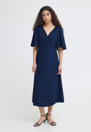 Naine, kellel on pikad lokkis juuksed, kannab tumesinist keskmise pikkusega kleiti liblikasvarrukatega ja musta kontsaga sandaale, seisab ühevärvilise taustaga.