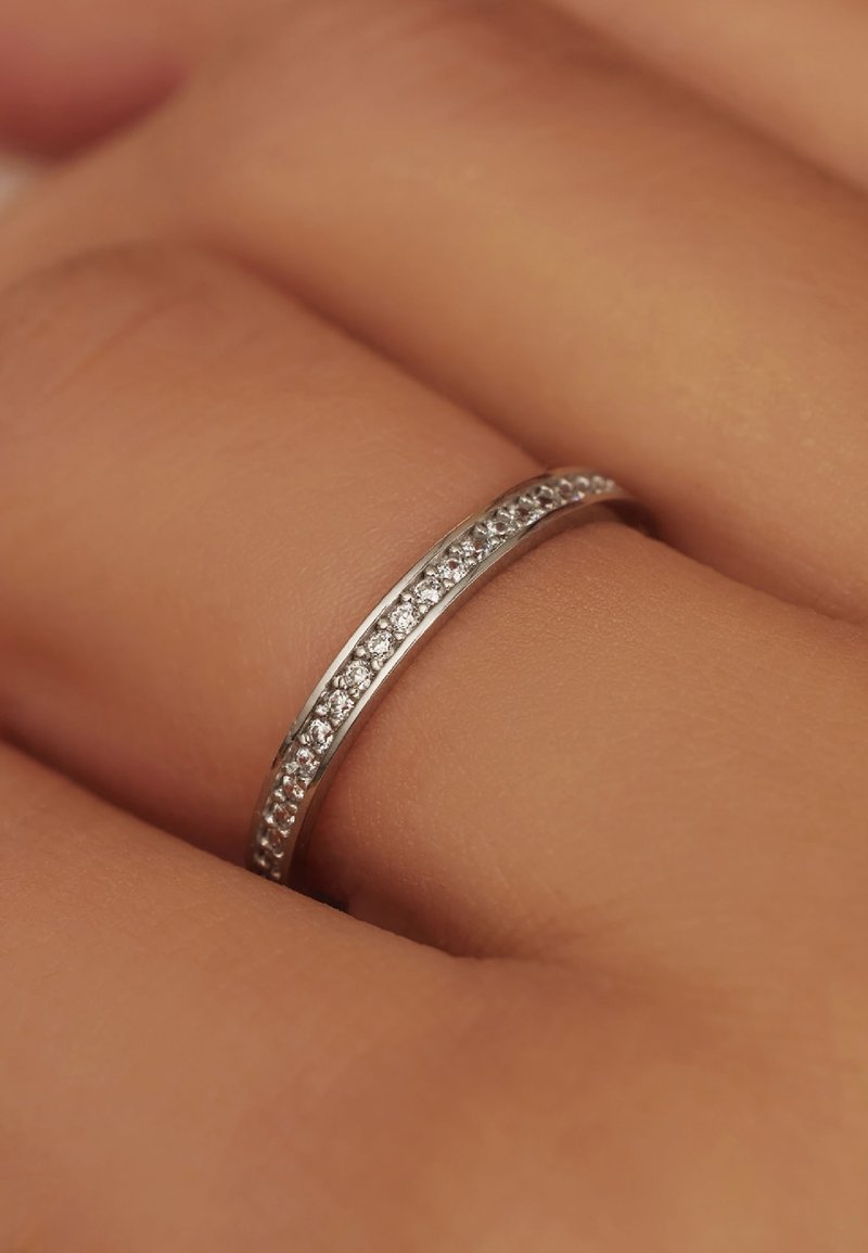 Bague en argent avec un anneau lisse, ornée d'une rangée de petites pierres étincelantes disposées sur le dessus. En arrière-plan, on aperçoit une main avec une teinte de peau.