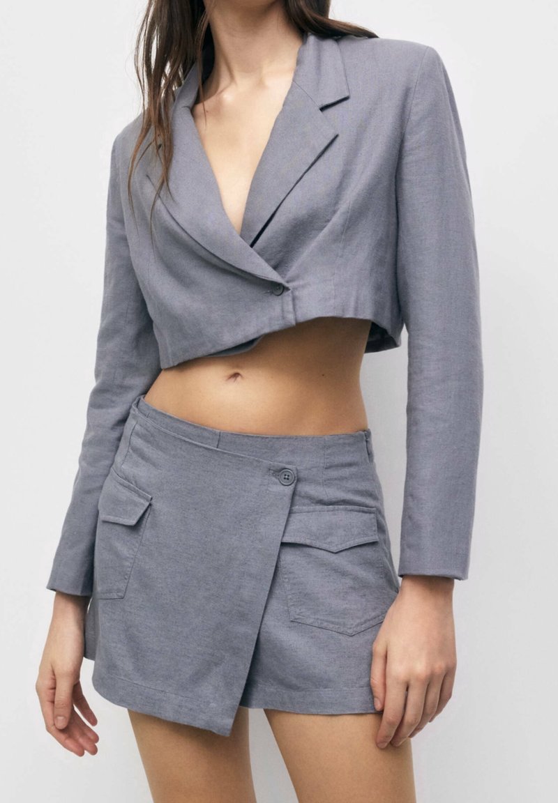 Blazer cropped in lino grigio con colletto a revers e maniche lunghe, abbinato a pantaloni shorts avvolgenti grigi con bottone e tasche laterali.