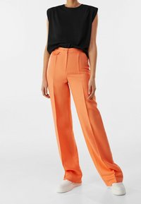 Top negro sin mangas combinado con pantalones anaranjados de pierna ancha de cintura alta. Los pantalones cuentan con un cierre de botón y una textura suave.