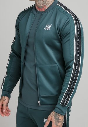 Homme portant un survêtement vert foncé avec le logo SikSilk et des bandes de marque sur les manches et le pantalon, montrant des tatouages sur le cou et les mains.