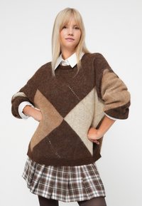 Blonde vrouw in een oversized bruin, beige en crème ruitjestrui over een wit overhemd en een geruiten rok, met haar handen op haar heupen, op een witte achtergrond.