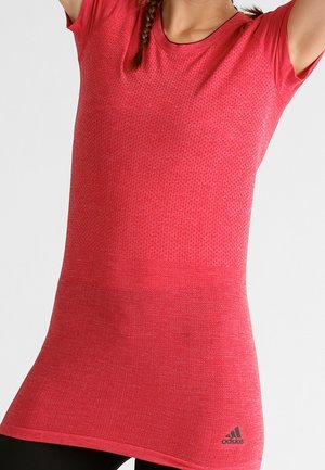 Frau trägt ein eng anliegendes, rot kurzärmeliges Sportshirt mit dezentem Muster und Adidas-Logo nahe dem Saum, hebt die Arme über den Kopf.