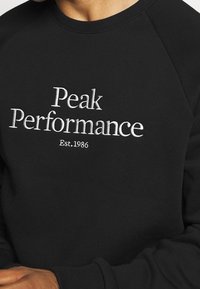 Sudadera negra con mangas raglán, que presenta el texto blanco bordado "Peak Performance" y "Est. 1986" en el frente.