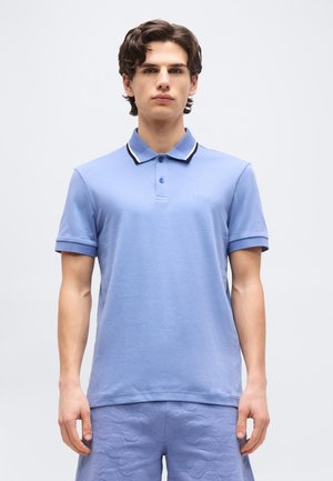 Jeune homme portant un polo bleu clair avec un col noir et blanc et un short assorti à motif, debout devant un fond uni.