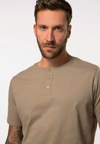 Jasnobrązowa bawełniana koszula henley z okrągłym dekoltem, krótkimi rękawami i czterema guzikami na plisie. Gładka tekstura, minimalistyczny design.