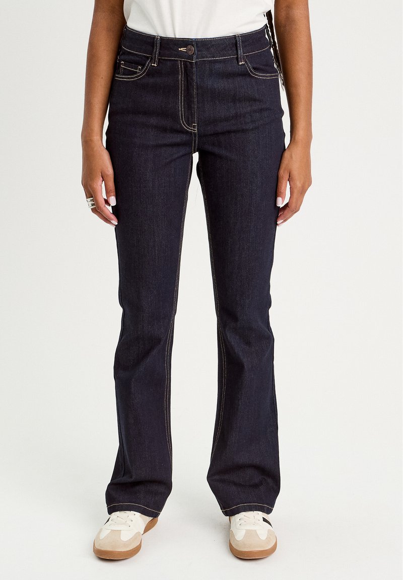 Dark blue denim jeans with a fitted waist, flared legs, and contrast stitching details. Paired with white sneakers.