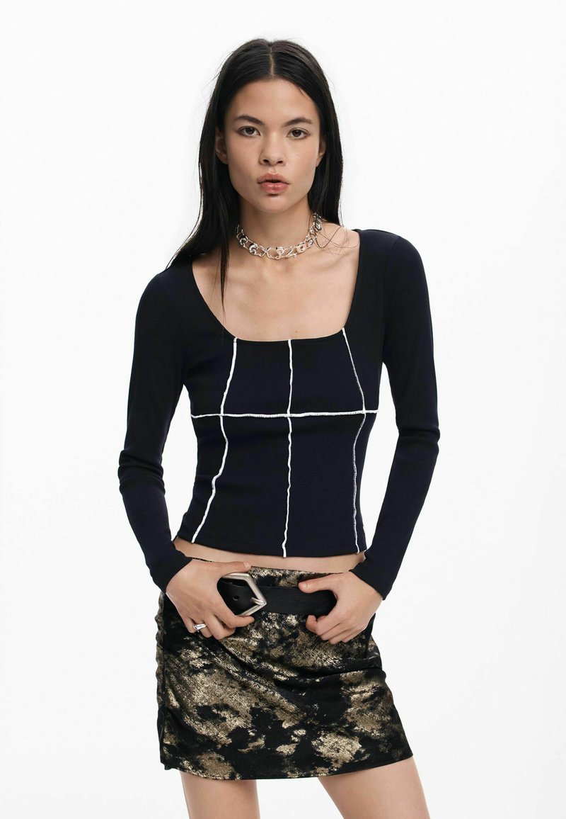 Mornarsko plavi rebrasti dukserica dugih rukava s bijelim geometrijskim šavovima, u kombinaciji s mini suknjom crne i zlatne boje s uzorkom te masivnom srebrnom ogrlicom.