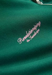 Grüner Sweatshirt-Stoff mit pinkem, geschwungenem Text, der "Revolutionizing the Soundscape" liest. Hat eine glatte Textur und einen Reißverschluss-Halsausschnitt.