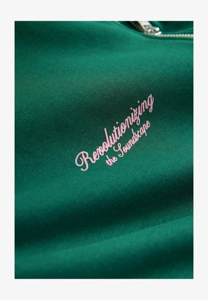 Zöld pulóver anyag, rózsaszín, írott betűkkel "Forradalmasítva a Hangeskét" felirattal. Simaság jellemzi, valamint cipzáras nyakkivágással rendelkezik.