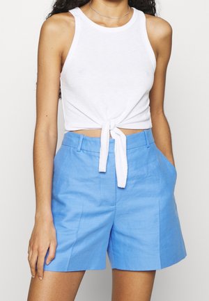 Kobieta ubrana w biały, bezrękawnik z wiązaniem z przodu oraz jasnoniebieskie, dopasowane szorty z wysokim stanem, z jedną ręką w kieszeni.
