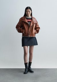 Brązowa skórzana kurtka z szerokim kołnierzem, zestawiona z pasiastym swetrem i dżinsową spódnicą. Modelka ma na sobie czarne botki za kostkę z prążkowanymi skarpetami.