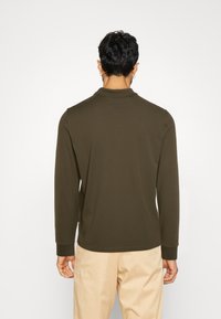 Olive green long-sleeve polo shirt with a collar and ribbed cuffs, made of a soft fabric; worn with light beige pants.