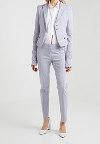 Žena v světle fialovém přiléhavém saku a odpovídajících úzkých kalhotách s bílou košilí a bílými vysokými podpatky na bílém pozadí.