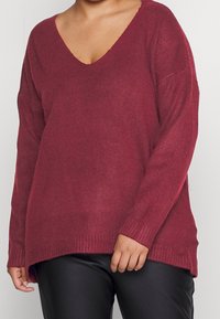 Burgunderfarbener Strickpullover mit V-Ausschnitt, langen Ärmeln und geripptem Saum. Hat einen lockeren Sitz und Seitenschlitze für zusätzliche Bewegungsfreiheit. Weicher Stoff.