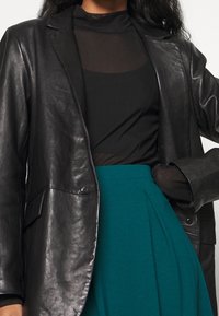 Svart läderblazer med en slät textur, ihopparad med en genomskinlig svart topp och en teal-färgad kjol med veck. Fokusera på strukturerad design och passform.