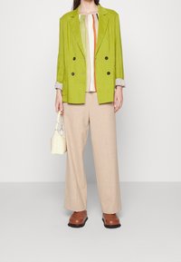 Donna con giacca doppiopetto verde lime, blusa a strisce multicolori, pantaloni beige a gamba larga, scarpe marroni, mentre tiene una piccola borsa bianca.