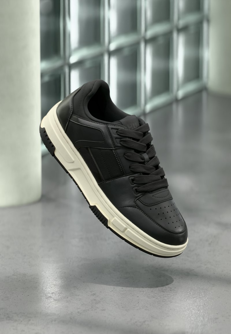 Baskets en cuir noir avec semelle blanche et lacets noirs flottant au-dessus d'un sol gris et d'un mur en blocs de verre en arrière-plan.