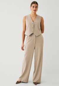 Beige Weste mit V-Ausschnitt und drei Knöpfen, kombiniert mit weiten beigen Hosen. Das Modell trägt Schuhe mit Tierdruck und Absatz. Glatte Textur des Stoffes.