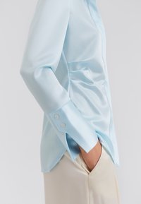 Γαλάζιο σατέν πουκάμισο με μακριά μανίκια, με μανσέτες με κουμπιά, εφαρμοστή γραμμή και λεία υφή, συνδυασμένο με κρεμ παντελόνια.