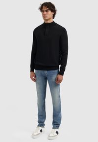 Mann in einem schwarzen langärmeligen Pullover mit einer Knopfleiste auf der Vorderseite, hellblauen, ausgewaschenen Jeans und weißen Sneakers mit schwarzen Details, stehend.