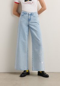 Hellblaue weite Jeans mit hoher Taille, die auf der Vorderseite bestickten Text aufweisen. Kombiniert mit schwarzen Loafers und gelben Socken.