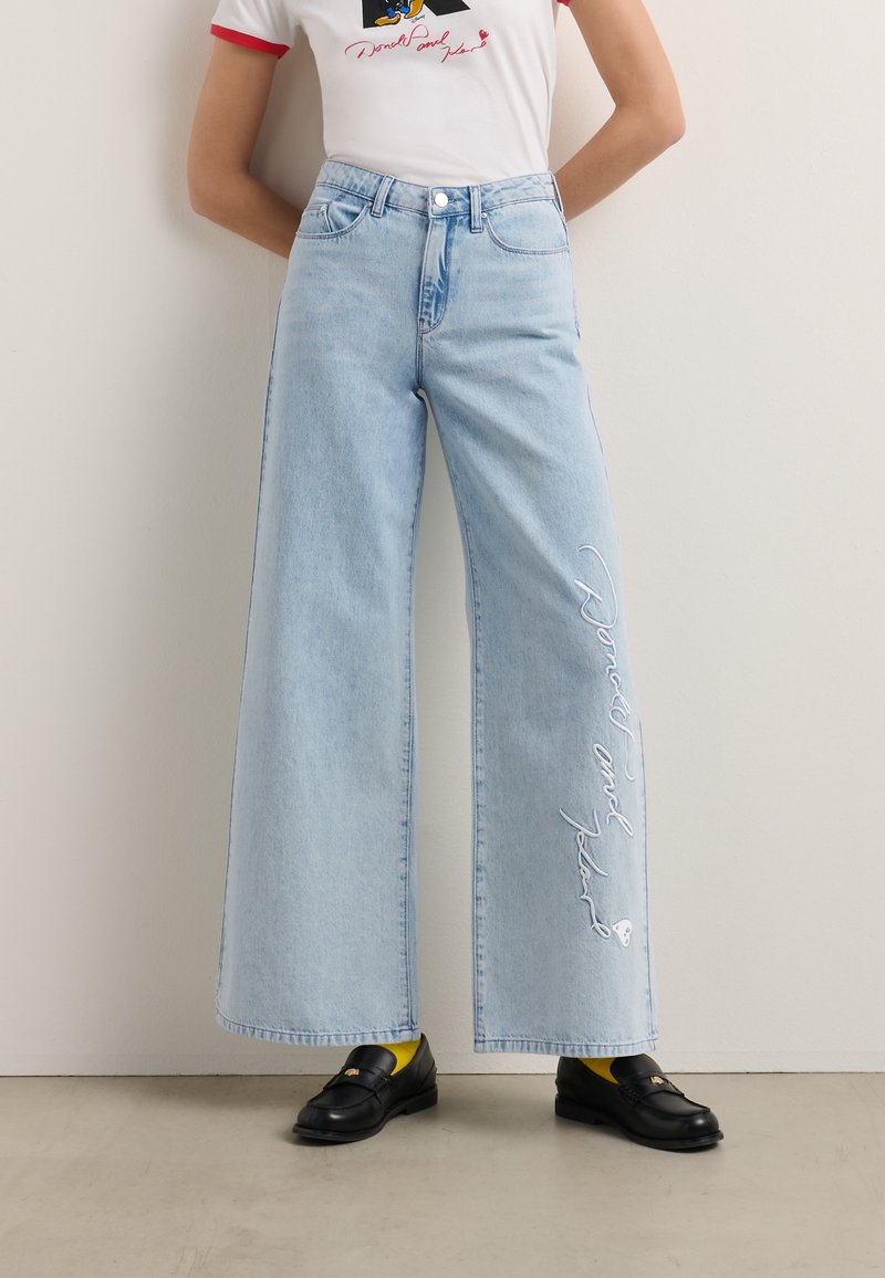 Hellblaue weite Jeans mit hoher Taille, die auf der Vorderseite bestickten Text aufweisen. Kombiniert mit schwarzen Loafers und gelben Socken.