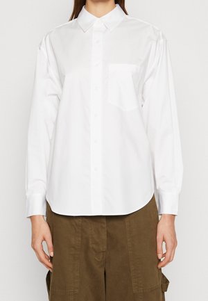 Personne portant une chemise blanche à manches longues, boutonnée, avec col et poche poitrine, associée à un pantalon marron.