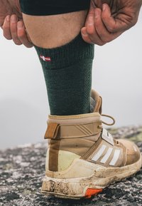 Grüner, gestrickter Socke mit weißen und roten Akzenten über einem beige, strukturierten Wanderschuh mit weißen Streifen und einer robusten Sohle, der auf felsigem Gelände ruht.