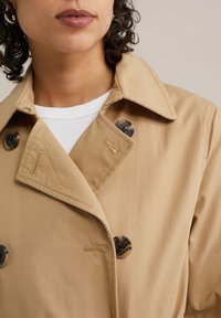 Beżowy płaszcz typu trencz z kołnierzem, ozdobiony dwoma dużymi okrągłymi guzikami. Tkanina jest gładka, z subtelnymi detalami przeszyć.