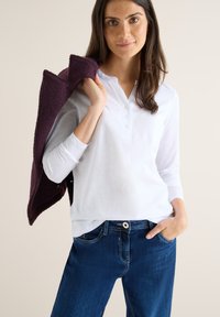 Hvid langærmet bluse med Henley-udskæring og knaplukning, parret med blå denimjeans og en mørk lilla, tekstureret sweater draperet over skulderen.