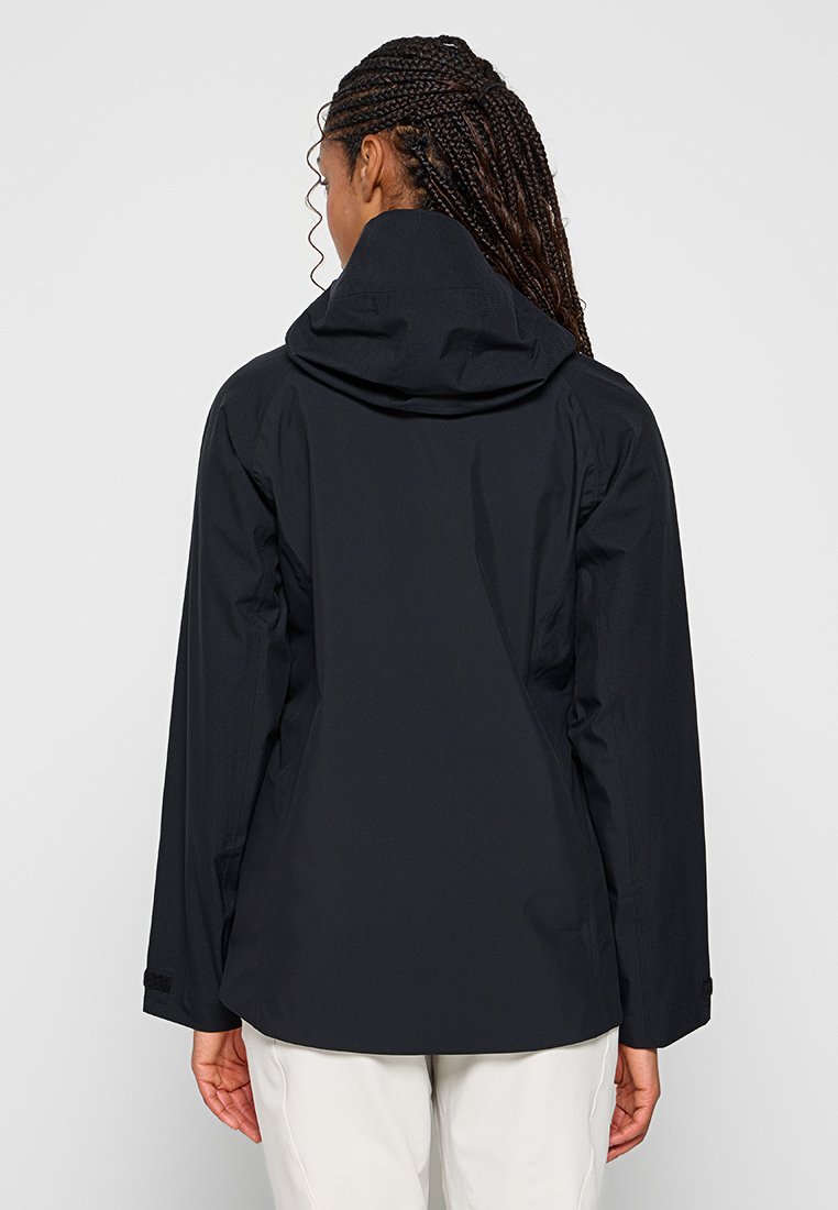 Kobieta z warkoczami, ubrana w czarną kurtkę z kapturem i jasne spodnie, pokazana od tyłu na jednolitym tle.