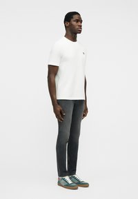 Hombre con camiseta blanca lisa de manga corta, jeans ajustados gris oscuro y zapatillas de ante verdes, de pie contra un fondo blanco.