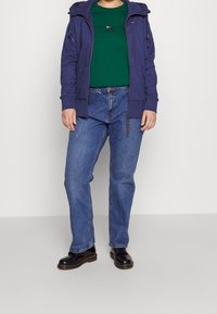 Marineblauwe rits hoodie over een groene t-shirt, gecombineerd met blauwe jeans en zwarte laarzen. De hoodie heeft twee voorzakken en een capuchon met trekkoord.