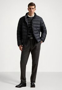 Svart dunjacka med hög krage och dragkedja, lager-på-lager över en mönstrad stickad tröja, kombinerad med grå byxor och svarta skor.