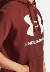 Bordowa bluza z kapturem z kieszenią typu kangur i dużym białym logo Under Armour na piersi. Gładka, miękka faktura materiału.
