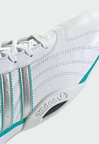 Weißer Ledersportschuh mit türkisblauen und silbernen Streifen, strukturierter Oberfläche und Gummisohle mit Logodetail an der Seite.