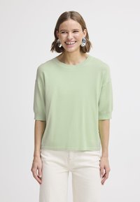 Maglione verde chiaro a maniche corte con vestibilità comoda, scollo tondo e texture morbida, abbinato a pantaloni color crema.