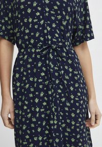 Marinblå klänning med grönt blommönster, korta ärmar och midjeband. Mjuk tyg med avslappnad passform, med små bladdetaljer överallt.