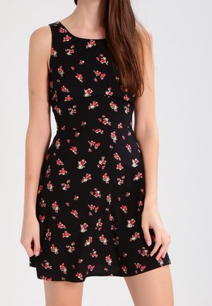 Mouwloze zwarte jurk met kleine rode en witte bloemmotieven, een getailleerde taille en een uitlopende rok, gedragen door een staand persoon.