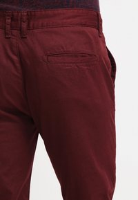 I pantaloni di cotone color bordeaux presentano una texture liscia, una vestibilità aderente e una tasca posteriore con contorno cucito.