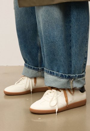 Des sneakers blanches avec des semelles en gomme et des lacets marron et blancs, portées avec un jean bleu ample retroussé sur un sol lisse.