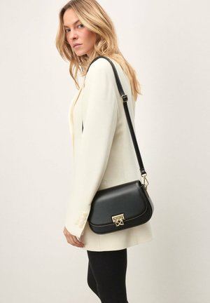 Lyshåret kvinde iført hvid blazer og sorte leggings, bærende på en sort lædertaske med guldlås.