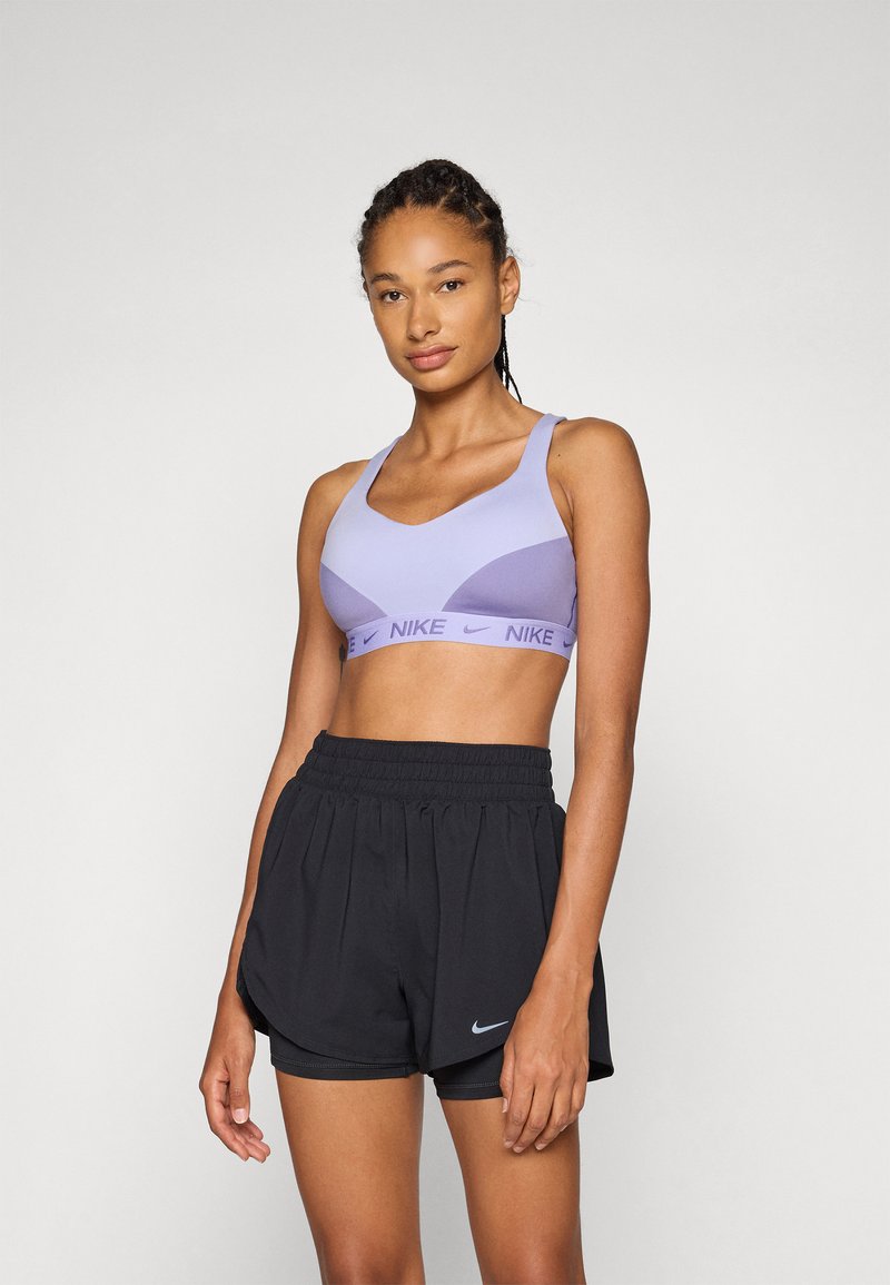 Lys lilla sports bra med v-halsdesign og brede stropper, med en Nike-logo på elastisk bånd. Kombinert med svarte løpeshorts med elastisk liv.