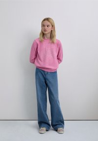 Light pink sweater with a skull and crossbones design, paired with wide-leg blue jeans and light-colored sneakers. Background is neutral.