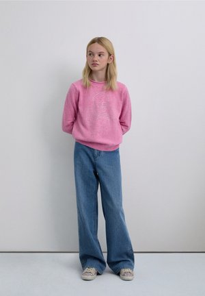 Vaaleanpunainen neulepaita, jossa pääkallo- ja luurankokuvio, yhdistetty leveälahkeisiin sinisiin farkkuihin ja vaaleanvärisiin tennareihin. Tausta on neutraali.