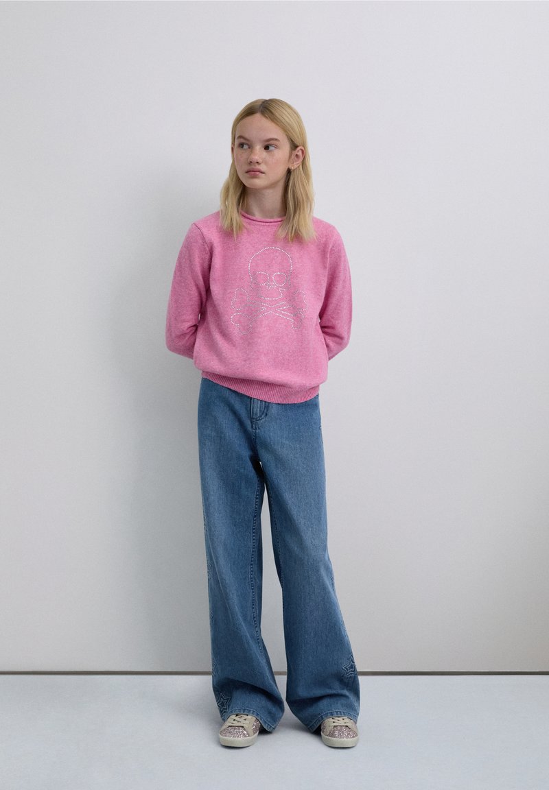 Light pink sweater with a skull and crossbones design, paired with wide-leg blue jeans and light-colored sneakers. Background is neutral.