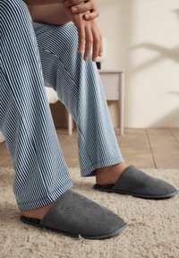Grau melierte Slipper ohne Riemen mit flacher Sohle, kombiniert mit blau-weißen gestreiften Schlafanzughosen, auf einem hellen Teppich platziert.