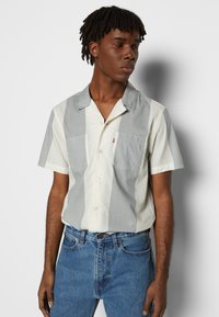 Camicia a maniche corte con bottoni di colore grigio chiaro e bianco sporco, dotata di due tasche sul petto, con colletto e tessuto leggermente strutturato.